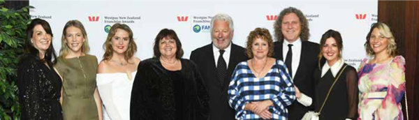 A group of individuals standing together at an awards event, dressed in formal attire. The background features a branded media wall with Westpac New Zealand Franchise Awards logos.