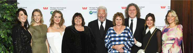 A group of individuals standing together at an awards event, dressed in formal attire. The background features a branded media wall with Westpac New Zealand Franchise Awards logos.